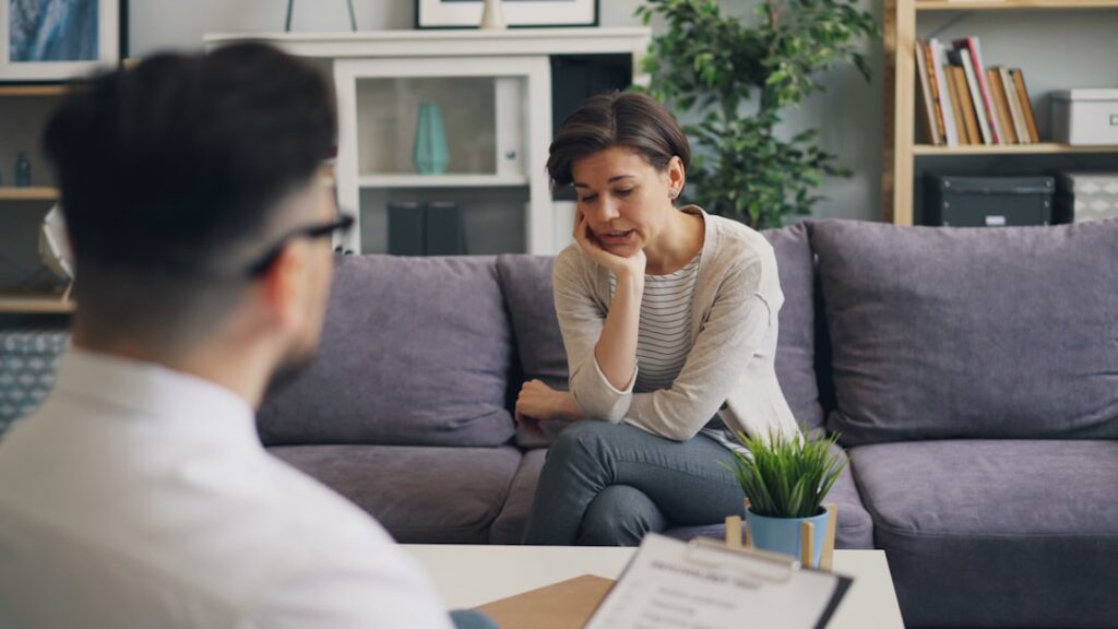 A counselor and client sitting in comfortable chairs, talking quietly in a softly lit room.