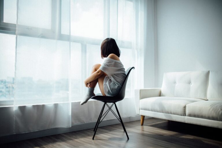 A person sits quietly by a window, looking outside on a gray day with a thoughtful expression.