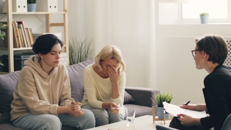 A couple sitting together on a home sofa, one partner offering comfort while the other looks down, hands intertwined.